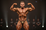 Thefreeway DN109 gold and black laminated competition bodybuilding posing trunks worn by a male bodybuilder on stage in a front double biceps pose