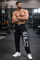 Unisex black bodybuilding training pants by Thefreeway Sportswear worn by a bodybuilder in the gym, front view with white leg logo